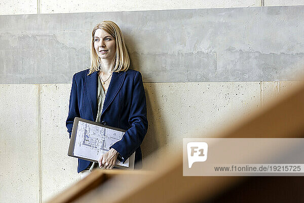 Thoughtful blond businesswoman standing with document and laptop in front of wall