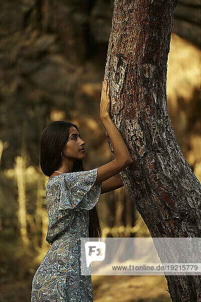 Teenage girl leaning on tree trunk in forest at sunset
