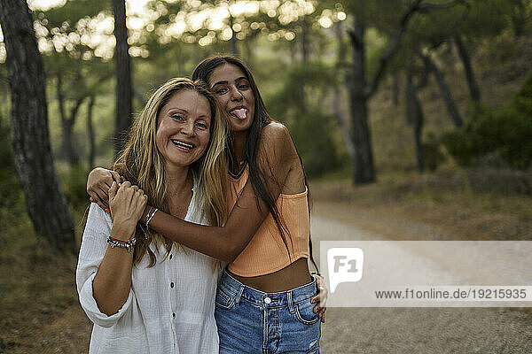 Happy teenage girl sticking tongue out hugging mother in forest