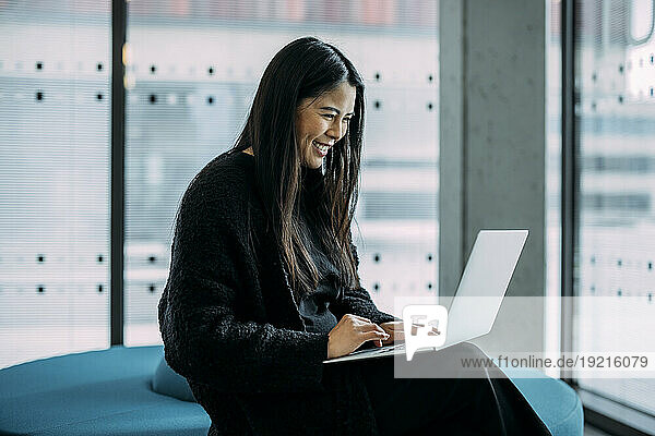 Smiling businesswoman using laptop on seat in office