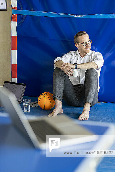 Smiling businessman sitting with coffee cup near basketball and laptop in office