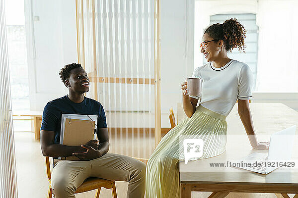 Happy young student with friend holding coffee cup and sitting on desk at university