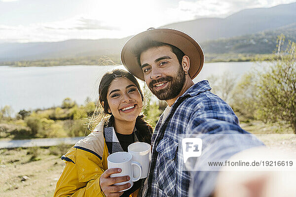 Smiling man taking selfie with girlfriend holding coffee mug