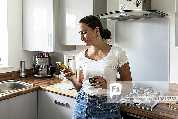 Smiling woman using smart phone and standing near kitchen at home
