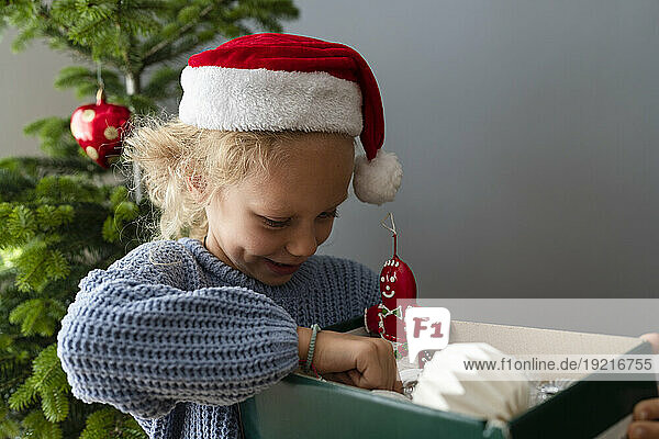 Girl wearing Santa hat holding box of Christmas decorations at home