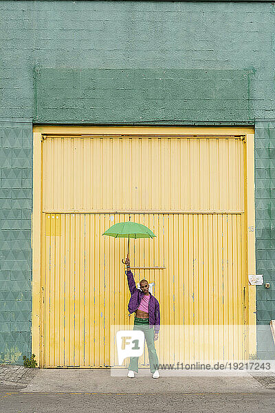 Non-binary person standing with umbrella in front of yellow shutter door