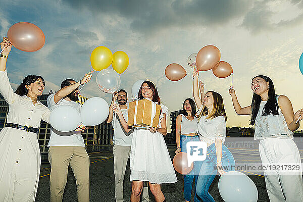 Cheerful friends celebrating woman's birthday with balloons and gift under cloudy sky