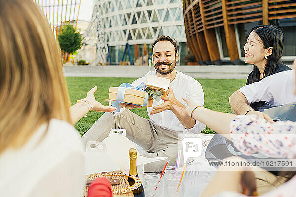 Friends giving gifts to happy man and enjoying picnic in park