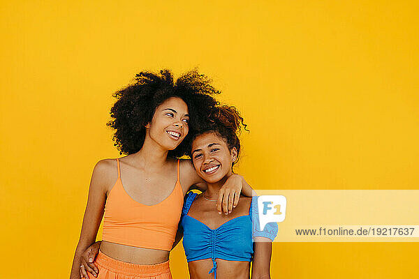Woman with arm around smiling friend against yellow background