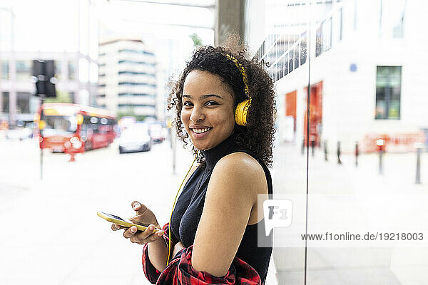 Happy woman holding smart phone and listening to music near building wall in city