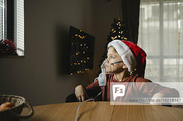 Thoughtful boy wearing santa hat using inhaler sitting at table