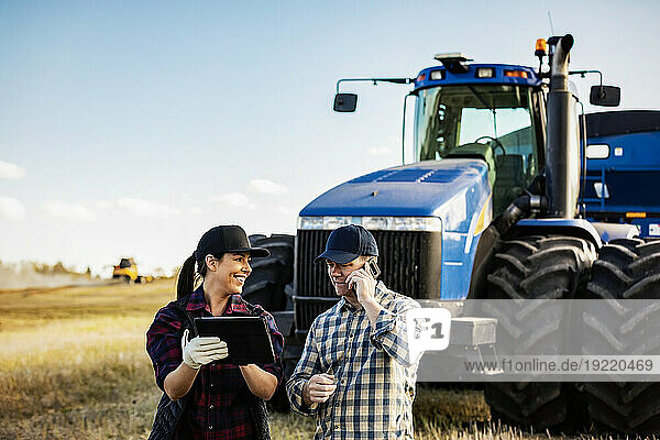 A husband and wife using portable wireless devices to manage and monitor their canola harvest and making a call while standing in front of a big tractor; Alcomdale  Alberta  Canada