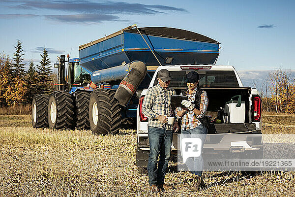 Husband and wife having a coffee while leaning on the tailgate of their half-ton truck and using a portable wireless device to manage and monitor their canola harvest with a semi-trailer grain hauler in the background; Alcomdale  Alberta  Canada