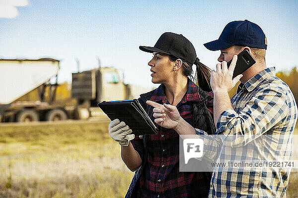 Close-up of a husband and wife using portable wireless devices to manage and monitor their canola harvest  making a call with a tractor trailer hauler in the background; Alcomdale  Alberta  Canada