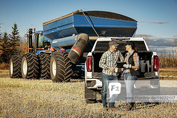 Husband and wife using a portable wireless device to manage and monitor their canola harvest and taking time for a coffee while leaning on the tailgate of their half-ton truck with a semi-trailer grain hauler in the background; Alcomdale  Alberta  Canada