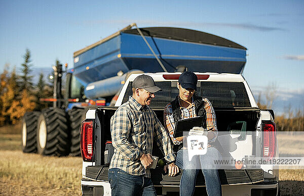 Husband and wife using a portable wireless device to manage and monitor their canola harvest and taking time for a coffee on the tailgate of their half-ton truck with a semi-trailer grain hauler in the background; Alcomdale  Alberta  Canada