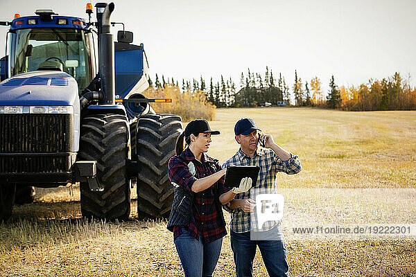 Husband and wife using portable wireless devices to manage and monitor their canola harvest and making a call while standing in front of a big tractor; Alcomdale  Alberta  Canada