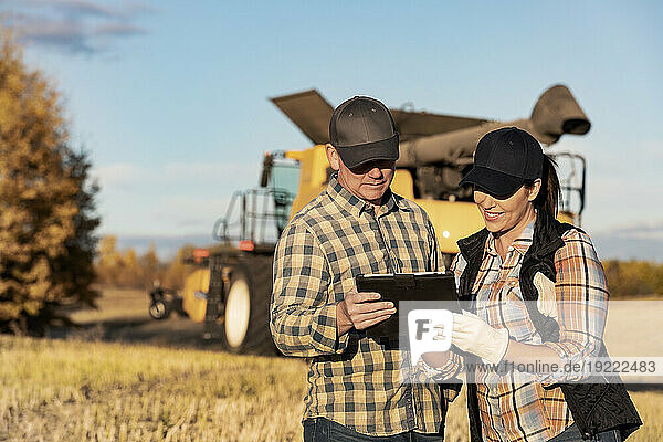 A husband and wife standing in front of a combine harvester and using a portable wireless device to manage and monitor the yield during their fall  canola harvest; Alcomdale  Alberta  Canada