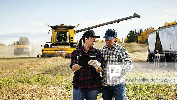 Husband and wife using portable wireless devices to manage and monitor their canola harvest while combine harvester offloads grain into a semi-trailer grain hauler in the background; Alcomdale  Alberta  Canada