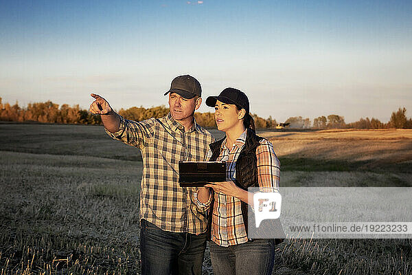A husband and wife standing in the fields at twilight using a portable wireless device to manage and monitor the yield during their fall  canola harvest; Alcomdale  Alberta  Canada