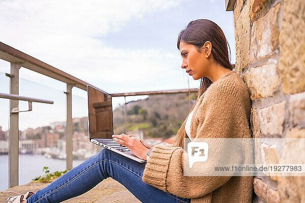 Eine junge lateinamerikanische Frau in einem weißen TShirt und einem braunen Wollpullover arbeitet auf der Terrasse ihres Hauses am Meer