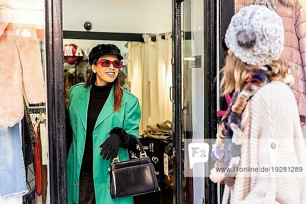 Junges kaukasisches Mädchen beim Einkaufen in der Stadt  begleitet von einer Verkäuferin  die sich das Schaufenster eines Bekleidungsgeschäfts ansieht. Sonnenbrille und rote Handschuhe