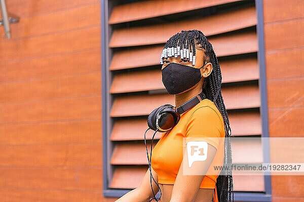 Schwarze Rasse Mädchen mit Maske von Coronavirus Pandemie  afrikanische ethnische Gruppe mit orangefarbenen Hemd in der Stadt. Cofing das Fahrrad in der Stadt geparkt