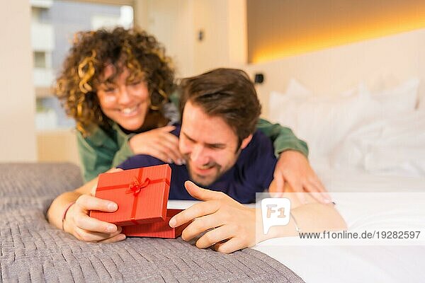 Valentinstag  ein junges kaukasisches Paar im Schlafanzug auf dem Bett  die Braut überrascht ihren Freund mit einem netten Geschenk