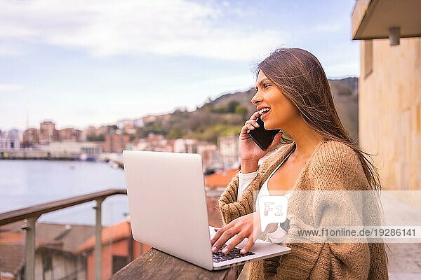 Eine junge lateinamerikanische Frau mit einem weißen TShirt und einem braunen Wollpullover  die auf der Terrasse eines Hauses am Meer telefoniert