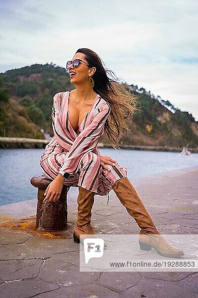 Porträt einer jungen lateinamerikanischen Frau in einem rosa Kleid  braunen Stiefeln und Sonnenbrille  die am Meer sitzend die Sommerferien genießt