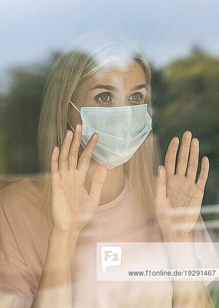 Frontansicht einer Frau mit medizinischer Maske  die während der Pandemie das Fenster berührt