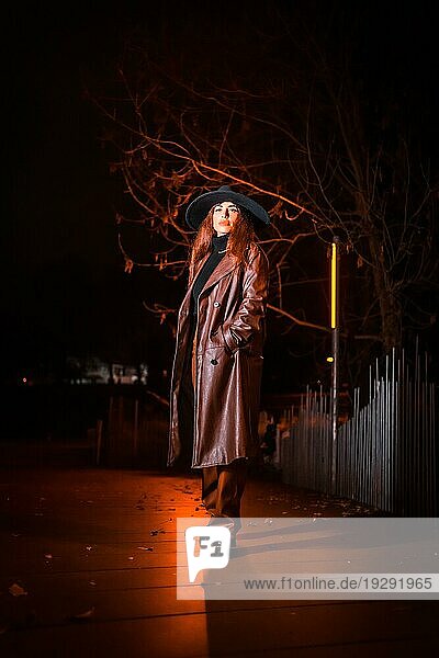 Mädchen mit Hut in einem Porträt bei Nacht in einem Regenmantel auf einem Paket. Kaukasisches brünettes Modell schaut in die Kamera
