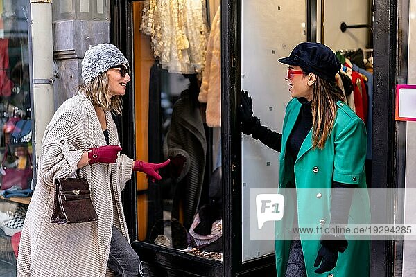 Junges blondes Mädchen beim Einkaufen in der Stadt  begleitet von einer Verkäuferin  die das Schaufenster eines Bekleidungsgeschäfts betrachtet. Sonnenbrille und rote Handschuhe