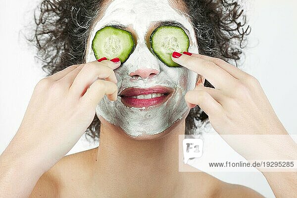 Lächelnde junge Frau mit Gesichtsmaske  die ihre Augen mit Gurkenscheiben bedeckt
