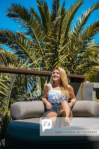 Sommerlicher Lebensstil einer jungen blonden kaukasischen Frau in einem weißen geblümten Top  die den Sommer auf einer Terrasse mit Palmen genießt. Sitzen auf einem Sofa auf einer Terrasse Sonnenbaden und Lächeln