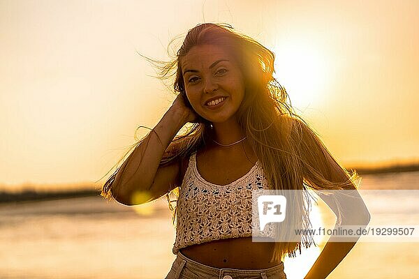 Sommerlicher Lebensstil. Eine junge blonde kaukasische Frau in einem weißen kurzen Wollpullover am Strand bei Sonnenuntergang. Lächelnd und Blick in die Kamera