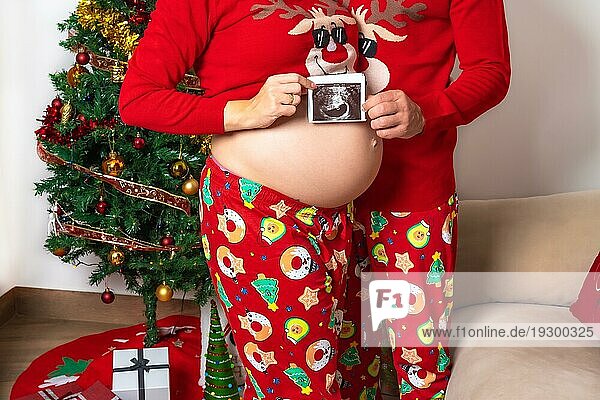 Junges Paar mit rotem Weihnachtsschmuck und Kleidung bei der Ultraschalluntersuchung des neuen Kindes für die Familie. Familie mit schwangerer Frau