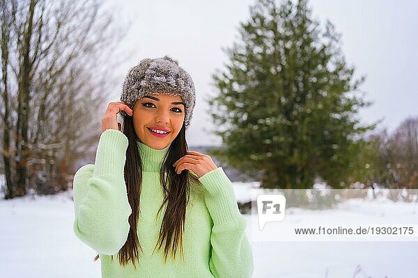 Winter Lifestyle  Porträt junge Brünette Kaukasierin mit grünem Outfit und Wollmütze  lächelnd im Schnee neben verschneiten Kiefern  Urlaub in der Natur