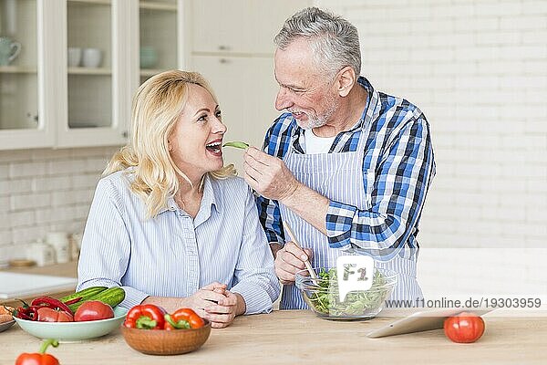 Lächelnder älterer Mann  der seiner Frau frischen grünen Salat in die Küche reicht