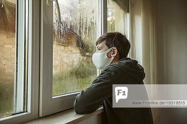 Seitlich stehender Junge mit Gesichtsmaske  der durch ein Fenster schaut
