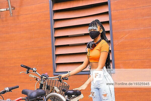 Schwarze Rasse Mädchen mit Maske von Coronavirus Pandemie  afrikanische ethnische Gruppe mit orangefarbenen Hemd in der Stadt. Cofing das Fahrrad in der Stadt geparkt