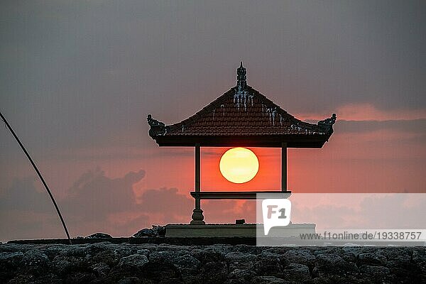 Sonnenaufgang am Sandstrand mit kleinen Tempeln im Wasser. Landschaftsaufnahme mit roter kreisförmiger Sonne am Strand von Sanur  Bali  Indonesien  Asien