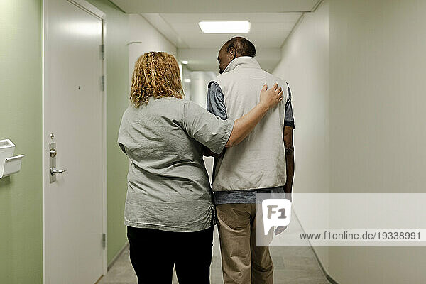 Rear view of female nurse walking with arm around senior man in corridor at retirement home