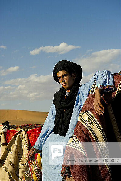 A desert guide in traditional dress stands in a tent camp with brightly colored blankets.