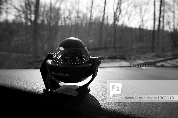 Black and white image of a compass on a dashboard with trees in the background.