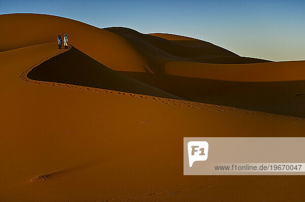 Two boys in traditional North African dress stand on the top of a sand dune at sunrise.