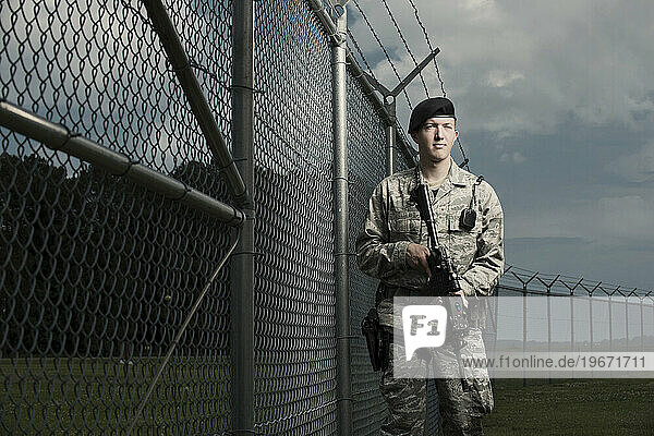 A Caucasian  male  Air Force Security Forces Airman in uniform poses with his M-4 rifle near the a secured fence line with barbed wire.