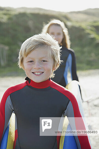 Smiling boy in a wetsuit carrying a surfboard with a girl