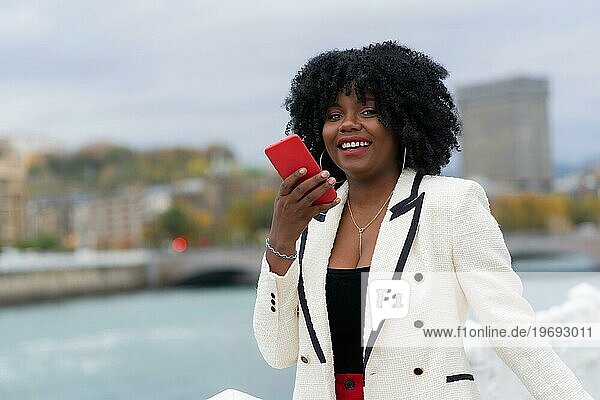 Elegante afroamerikanische Geschäftsfrau  die eine Sprachnachricht im Freien neben einem städtischen Fluss sendet