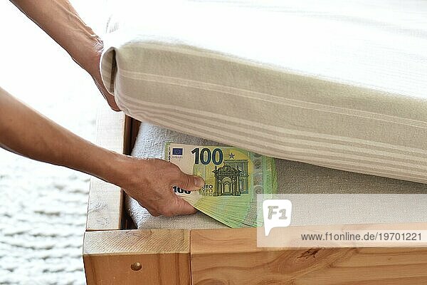 Eine Hand deponiert viele einhundert Euro Geldscheine unter einer Matratze  Symbolbild für Sparen in Bargeld